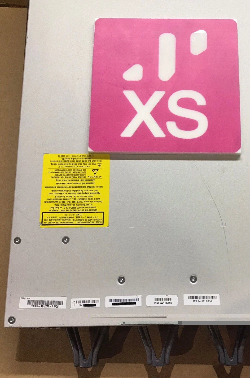 Pink rectangular sticker with white xs logo and stylized signal bars on metallic gray cisco catalyst 9300 48-port switch